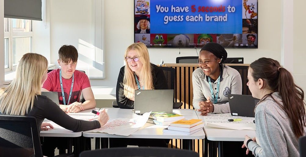 5 Business students sitting in a well lit room around a table smiling